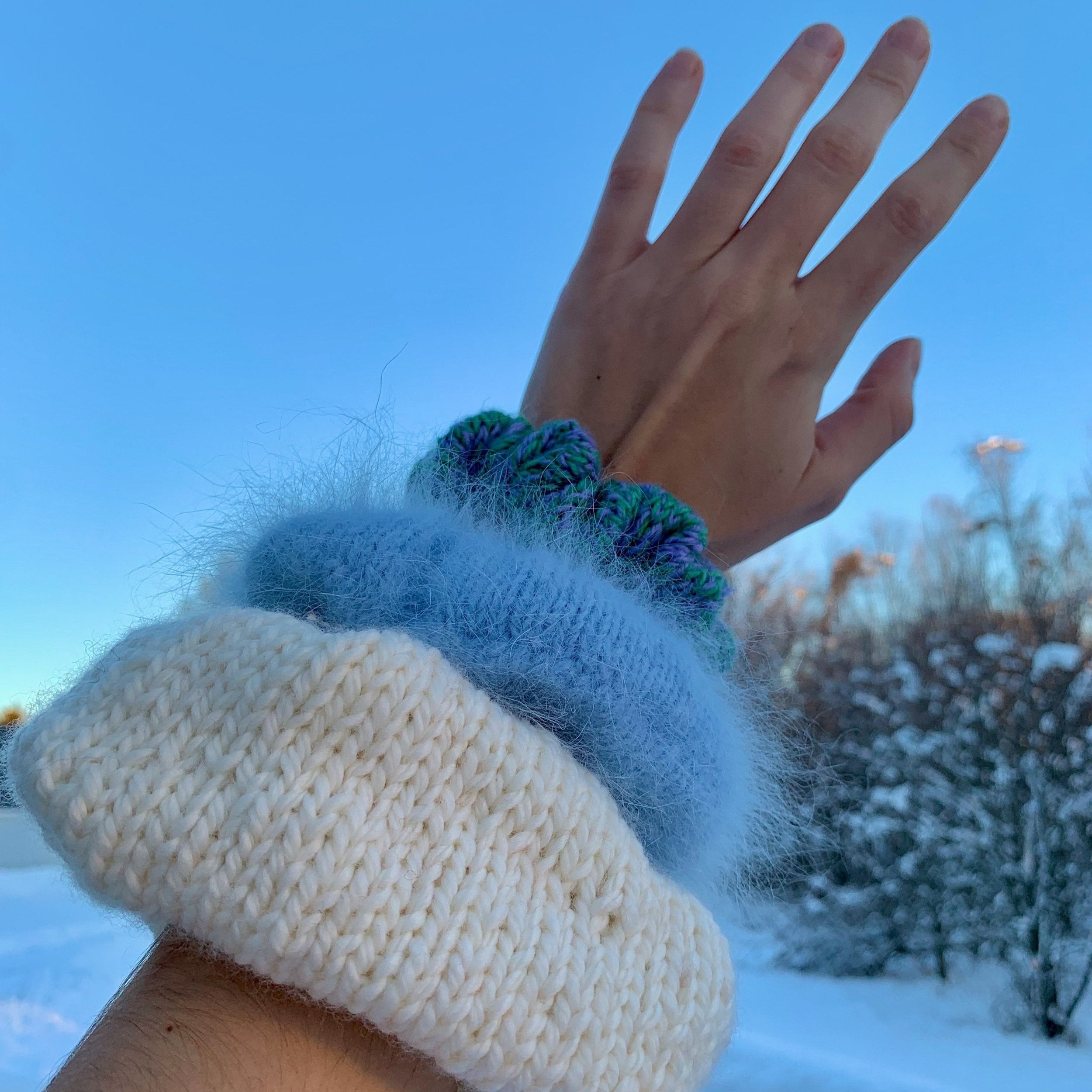 A person holding their left hand up showcasing three different sized knitted scrunchies on the arm