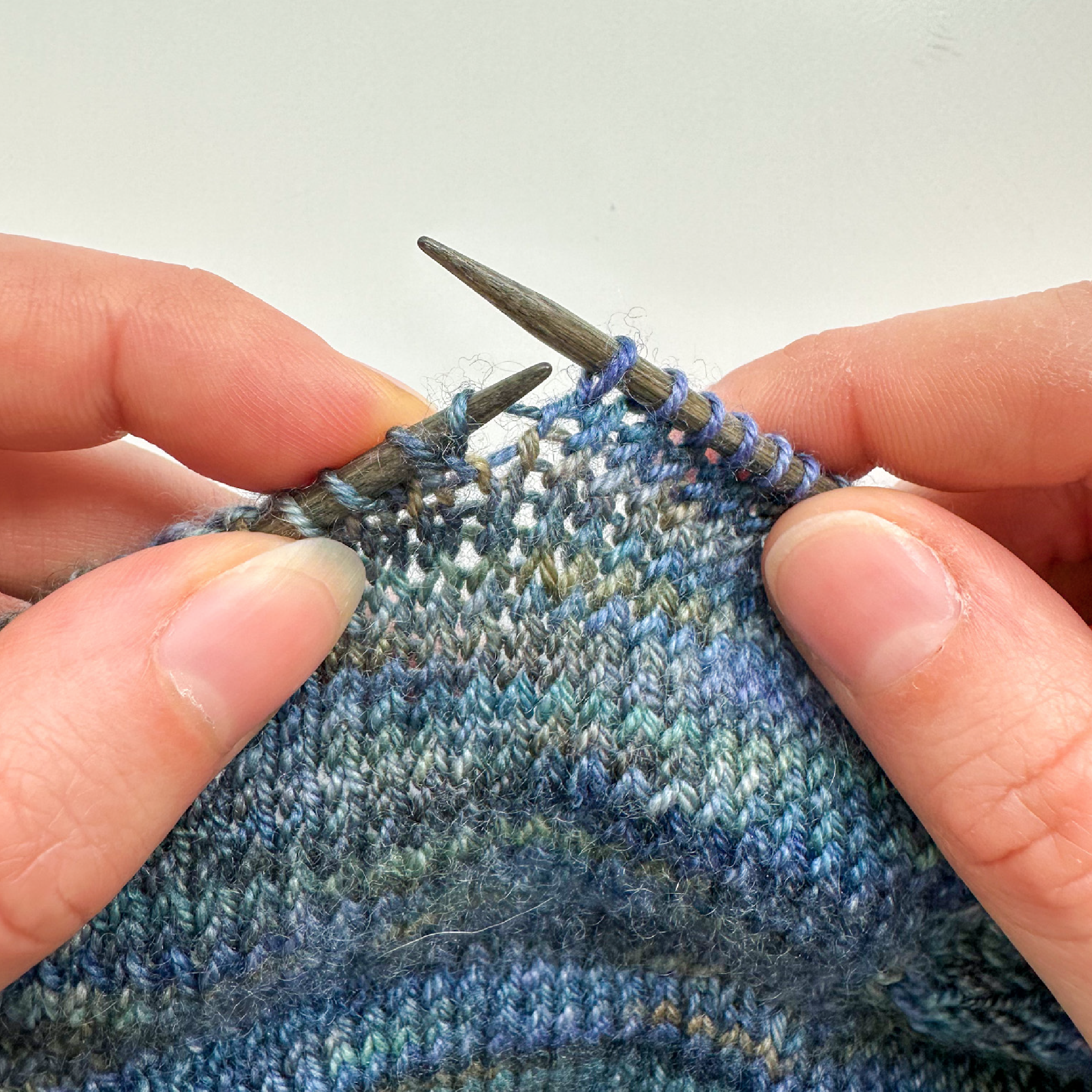 Close-up of hands knitting with two needles, showing the process of creating a M1L or M1R increase in a variegated blue yarn