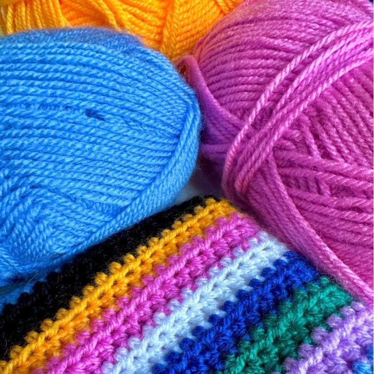Close-up of colorful acrylic yarn balls in blue, yellow, and pink, with a partially crocheted piece featuring various bright stripes.