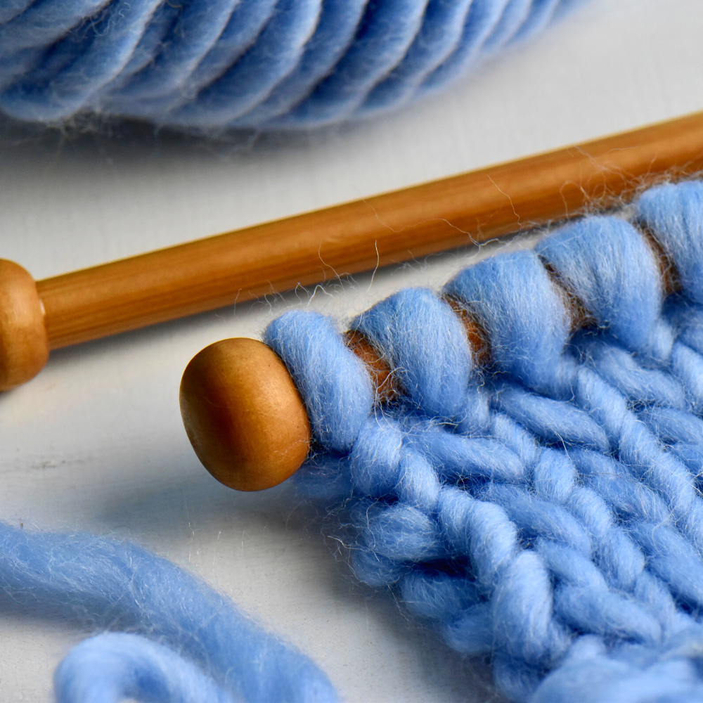Close-up of chunky blue yarn being knitted on a wooden needle.