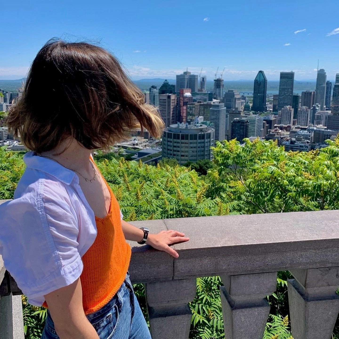 A woman in a knit orange top and jeans gazes out over a vibrant city skyline.