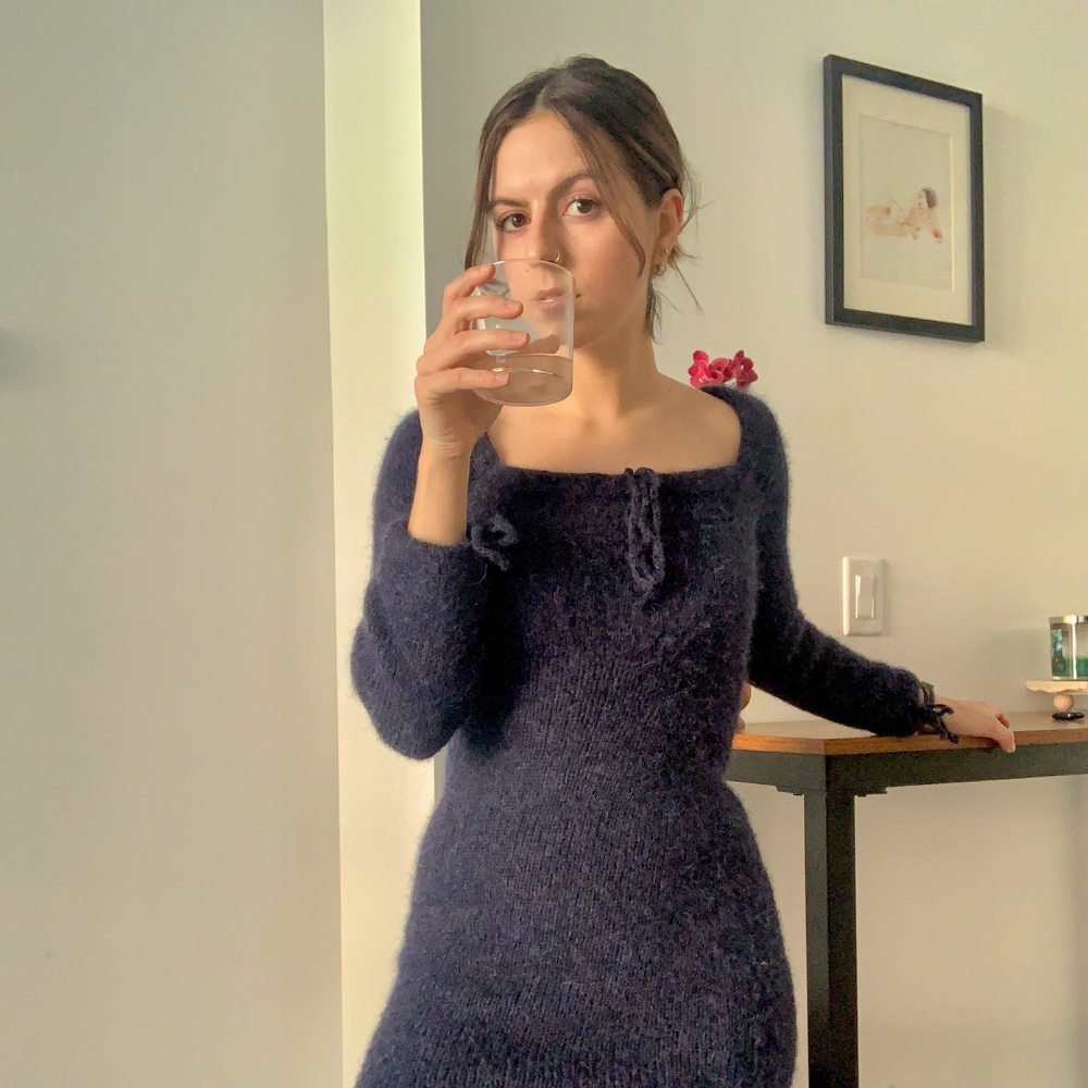 A woman wearing a blue knitted dress holds a glass of water, highlighting the beauty of sustainable fashion and fiber arts.