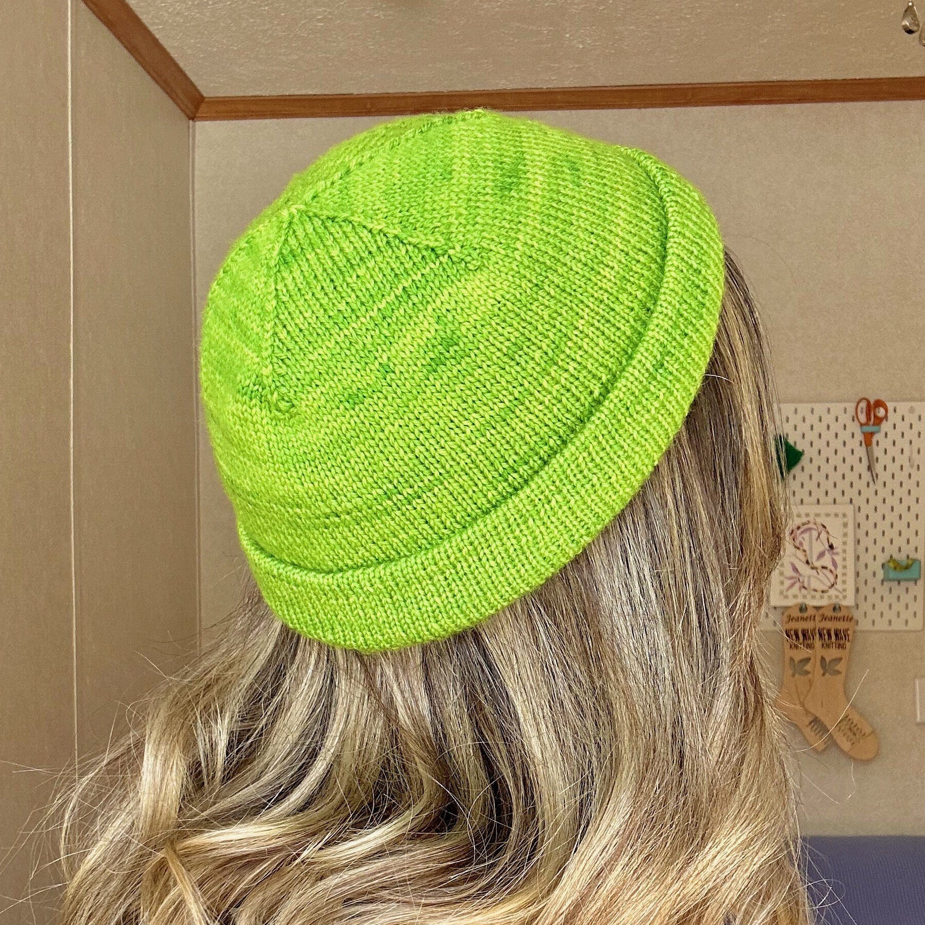 A back view of a woman in a green knitted hat showcases the Short Staple Beanie knitting pattern