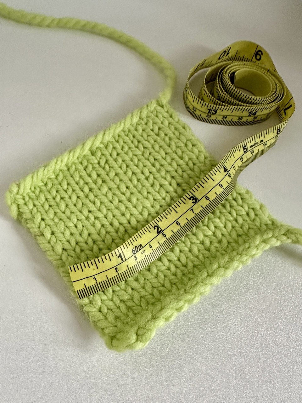 A lime green knitted gauge swatch being measured with a yellow tape measure, emphasizing the importance of accurate gauge in knitting projects.