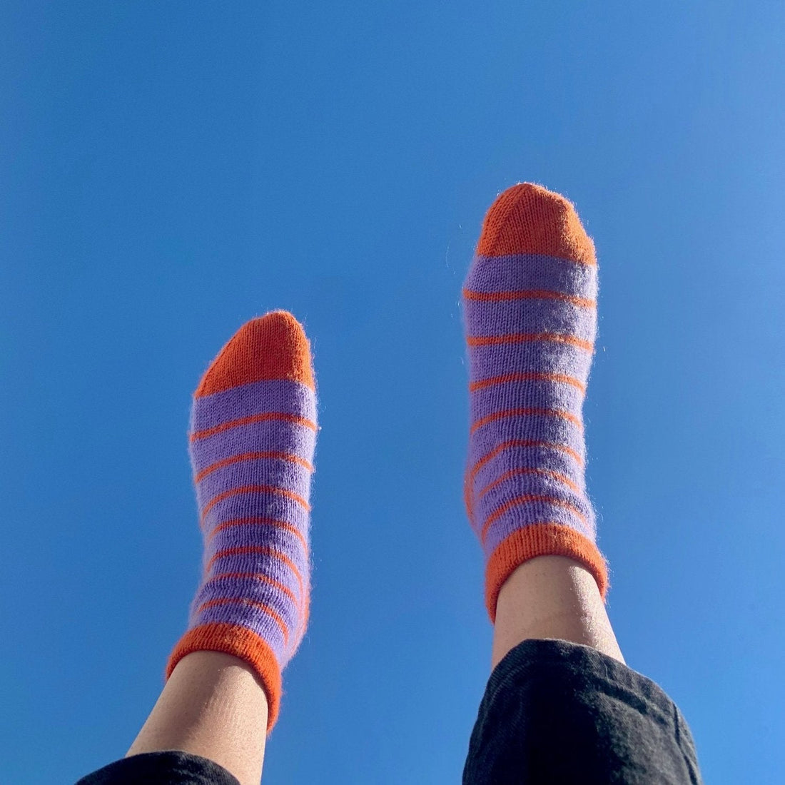 A person’s feet in the air, showcasing vibrant purple and orange socks made with the Staple Sock Pattern