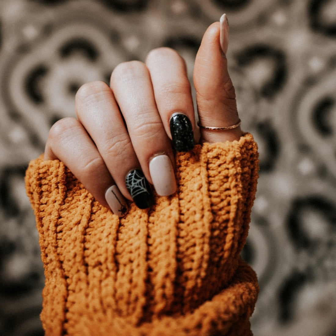A hand with manicured nails, featuring a spider web design, is shown against a patterned background. The hand is partially covered by an orange knit sleeve.