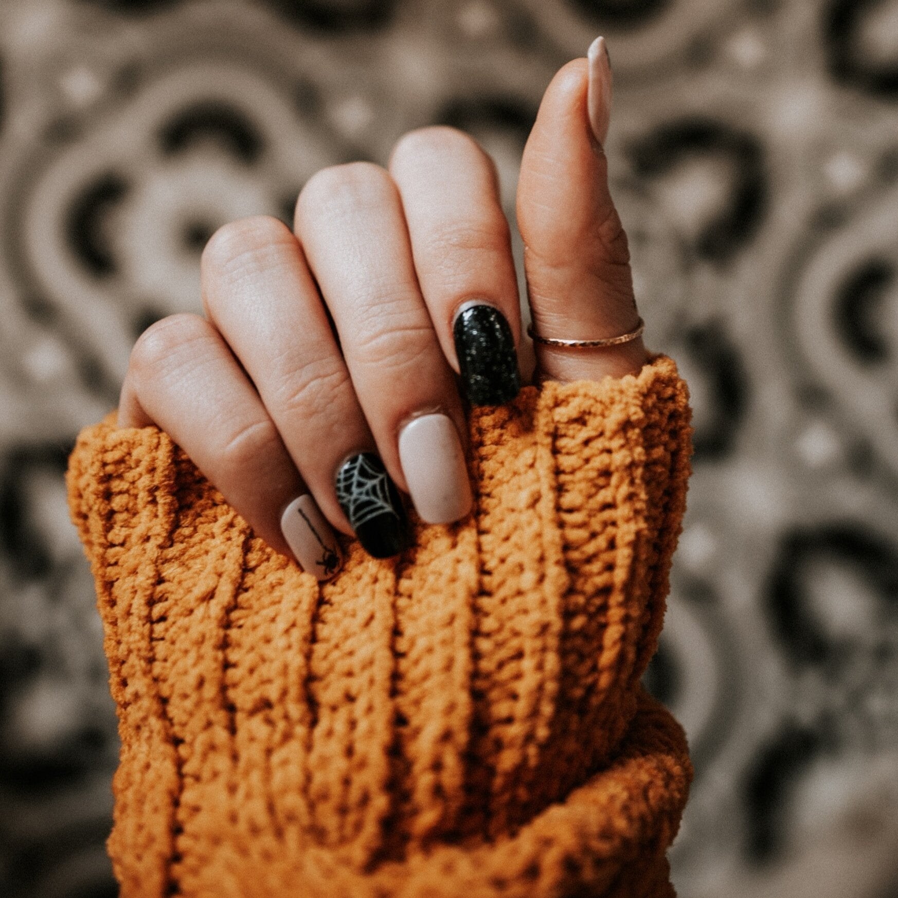 A hand with manicured nails, featuring a spider web design, is shown against a patterned background. The hand is partially covered by an orange knit sleeve.