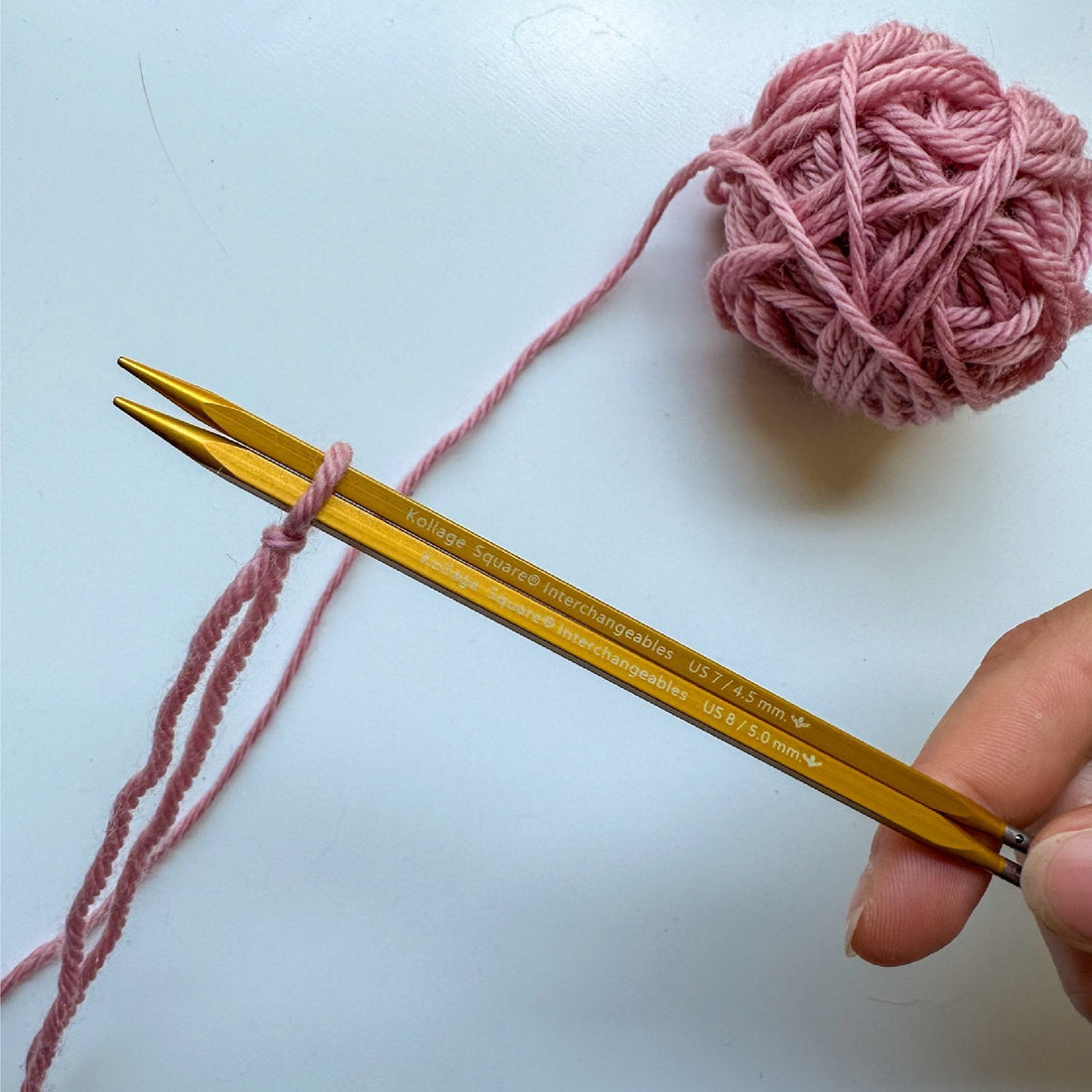 A hand holding two square knitting needles with pink yarn looped around them and a pink ball of yarn on the side