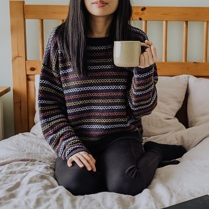 a person wearing a knitted sweater sitting on a bed holding a cup 