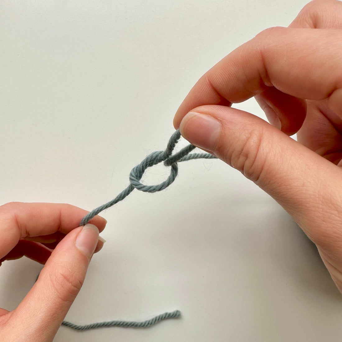 Two hands grasp a small piece of yarn, demonstrating the technique of creating a slip knot for knitting