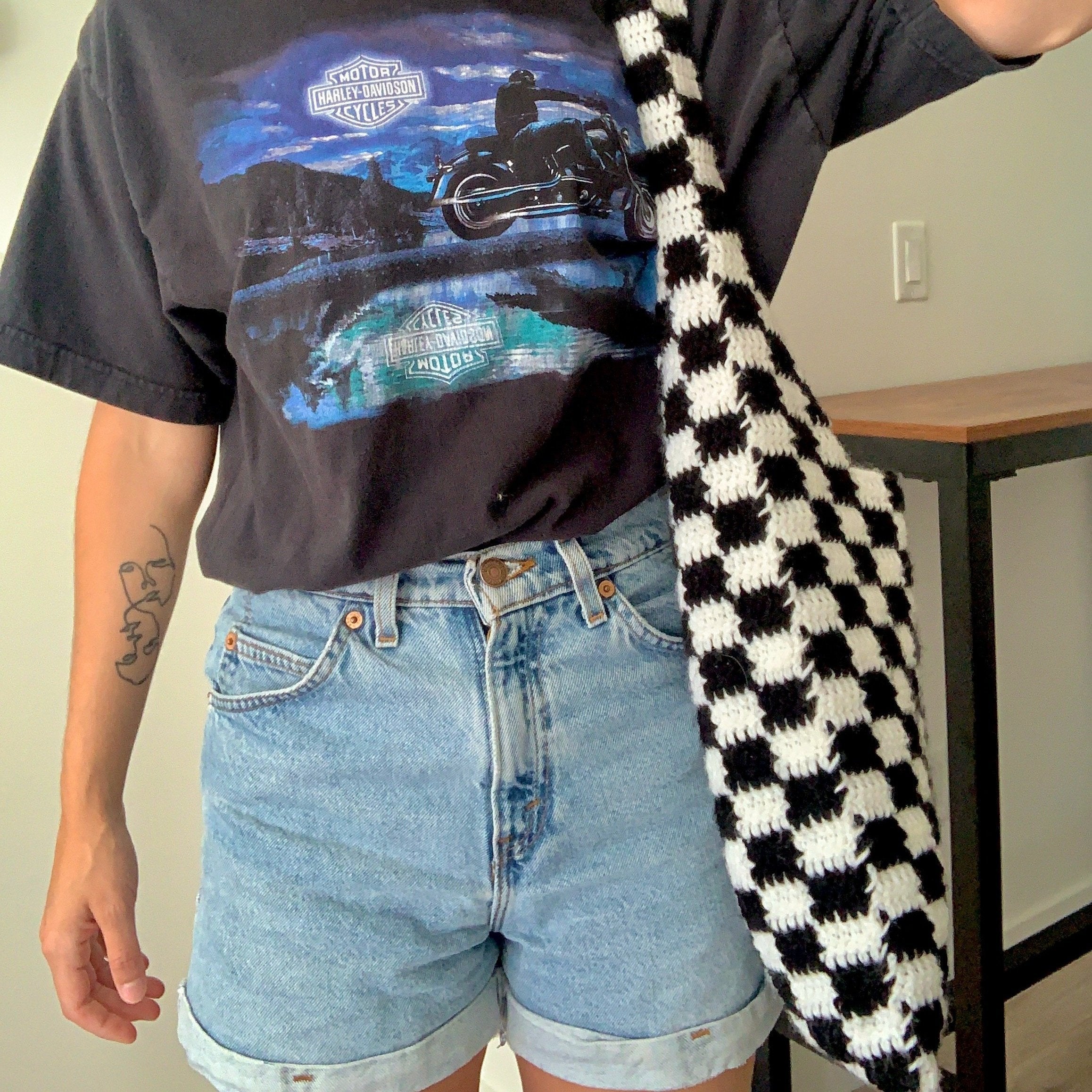 A woman in a shirt and shorts stands confidently, showcasing a checkered crochet bag