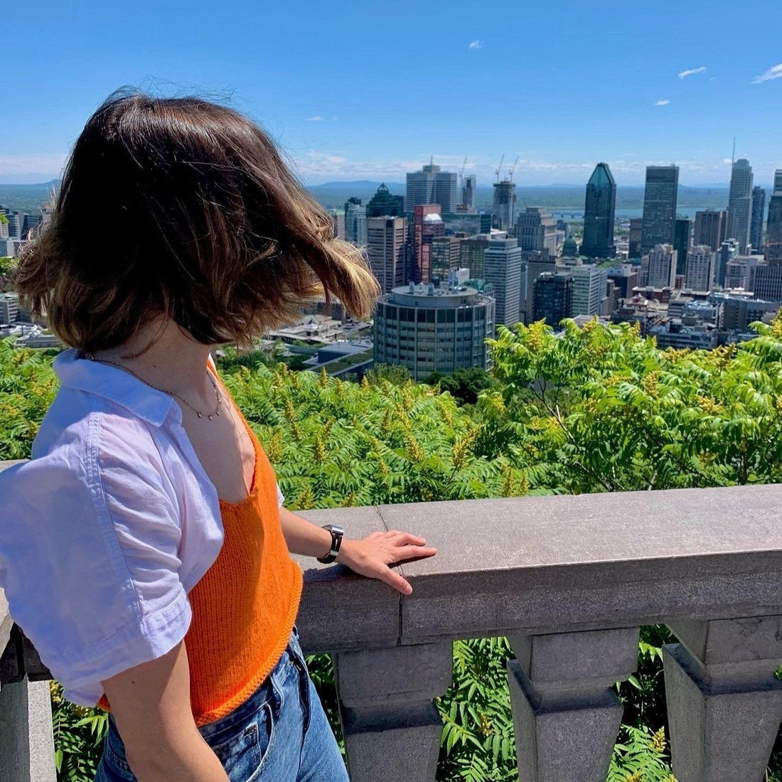 A woman in a knit orange top and jeans gazes out over a vibrant city skyline.