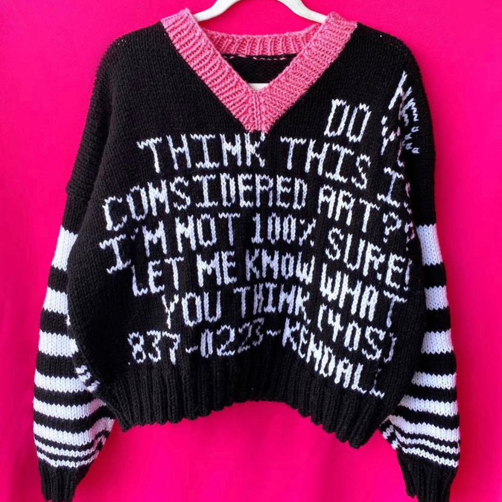 A pink knitted sweater displaying the text "think this is considered art?," exploring the dialogue between craft and artistic expression.