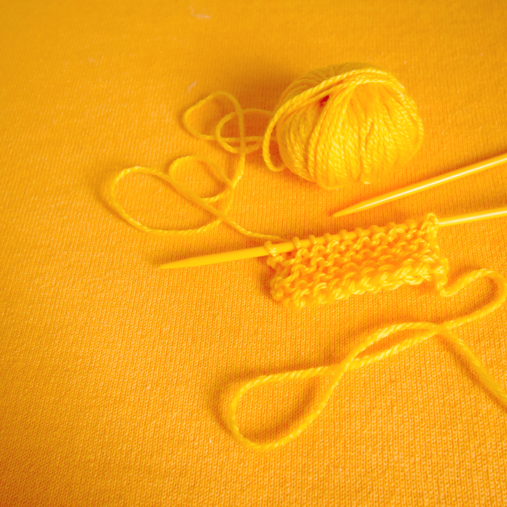 A vibrant yellow ball of yarn accompanied by two knitting needles, against a yellow knitted background, showcasing a new knitting project.