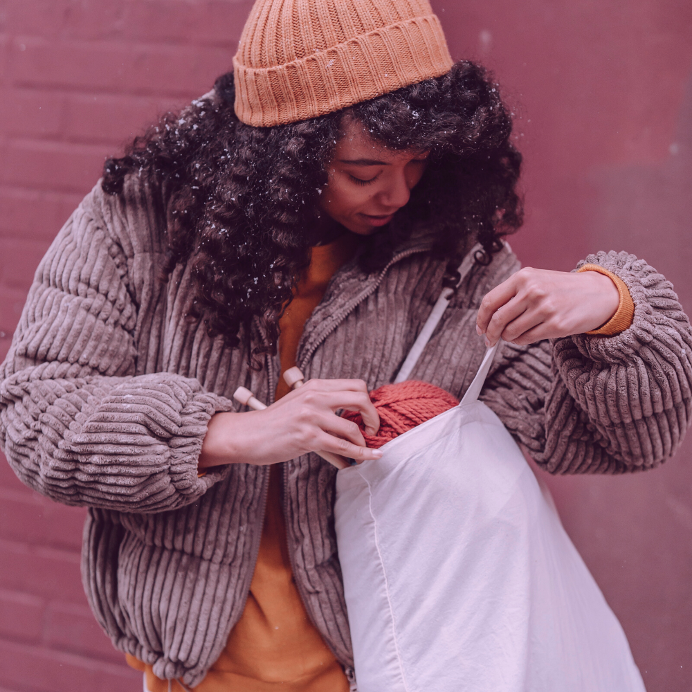 A woman with curly hair wearing a knitted hat while holding a bag full of knitting supplies celebrating World Wide Knit in Public Day