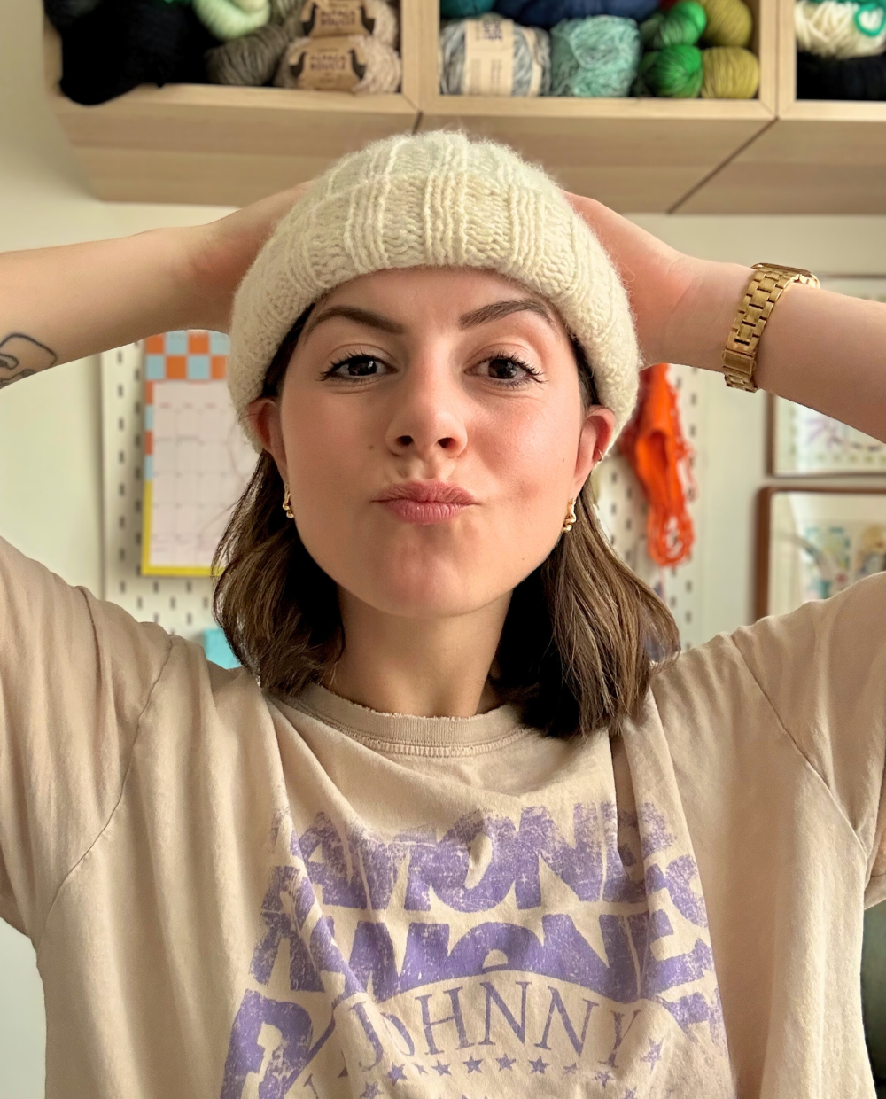 Person wearing a beige knit beanie and t-shirt with text, standing in front of a shelf with yarn.